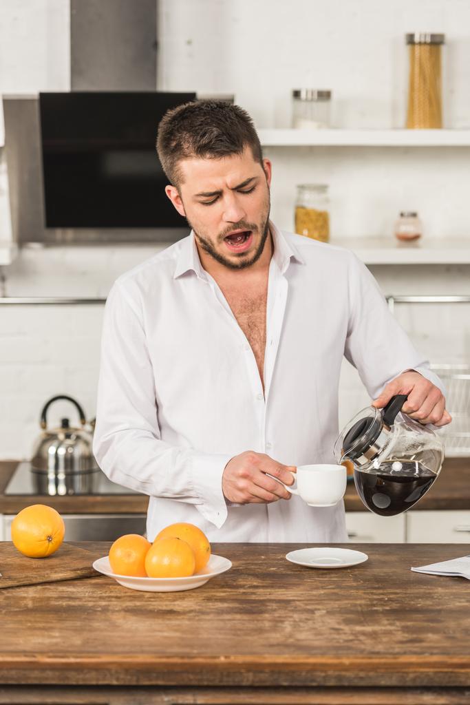 όμορφος άνδρας χασμουρητό και ρίχνει καφέ στο φλιτζάνι πρωί στην κουζίνα - Φωτογραφία, εικόνα