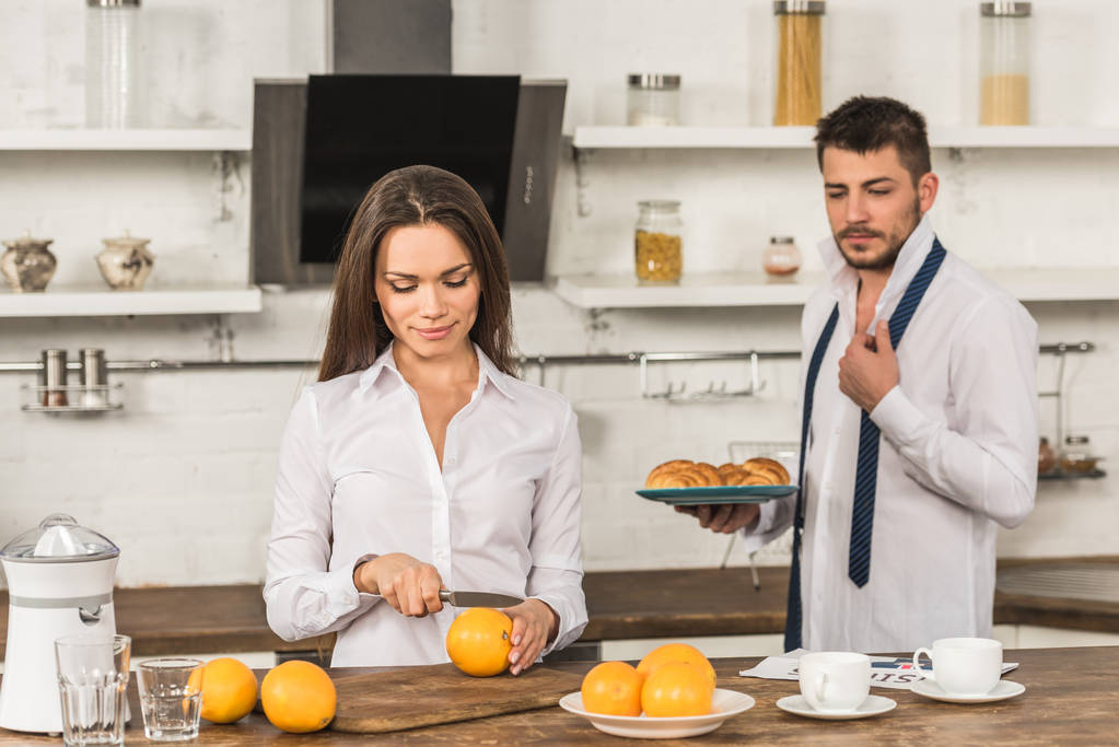 парень держит тарелку с круассанами и подружка режет апельсины дома
 - Фото, изображение