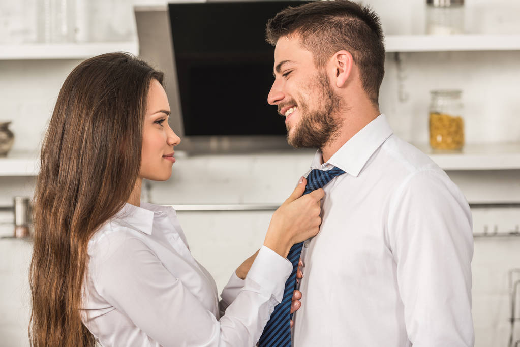 portrait of girlfriend tying smiling boyfriend tie in morning at kitchen, sexism concept - Photo, Image