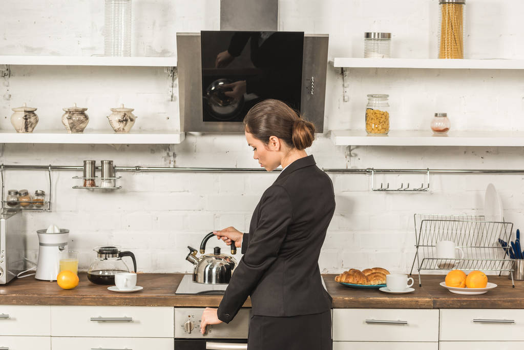 mulher atraente em terno colocando chaleira no fogão de manhã na cozinha
 - Foto, Imagem