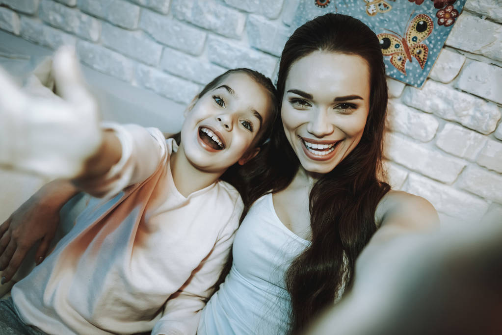 Moeder met dochter zit op de Bank. Mensen doen een Selfie op mobiele telefoon. Moeder met dochter is gelukkig en Smiling. Muur met foto's op de achtergrond. 'S avonds de tijd. Interieur. - Foto, afbeelding