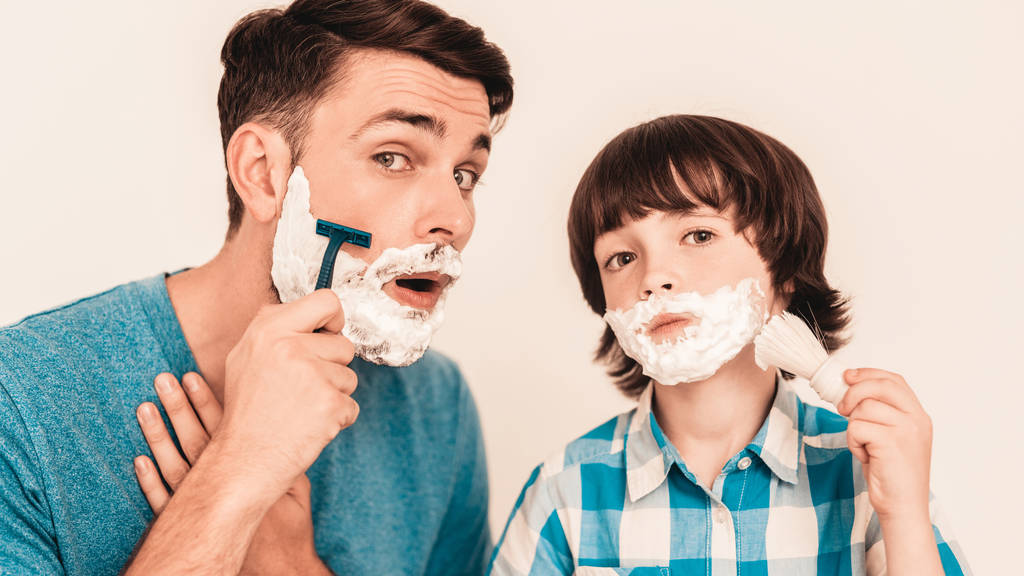 若い父は、家でひげをそるの幼い息子を教えています。顔に泡。シェービング ツールです。かみそりの刃で。幸せな家族の概念。シャツの少年。衛生とスキンケア概念。衛生的な石鹸. - 写真・画像