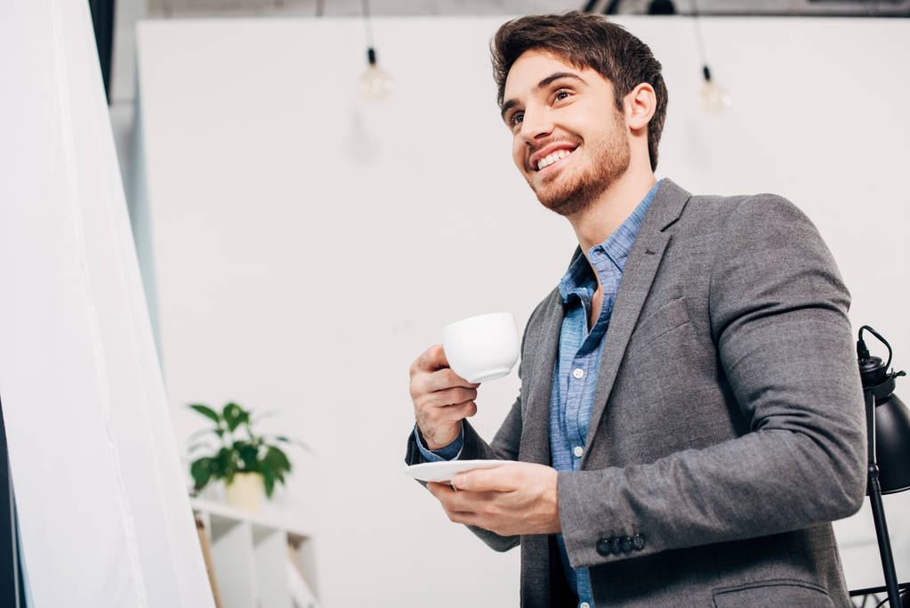 handsome office manager smiling and holding coffee cup in office - Photo, Image
