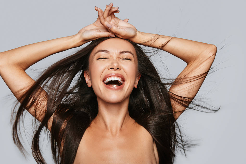 laughing young Asian woman keeping eyes closed and standing against grey background shirtless - Photo, Image