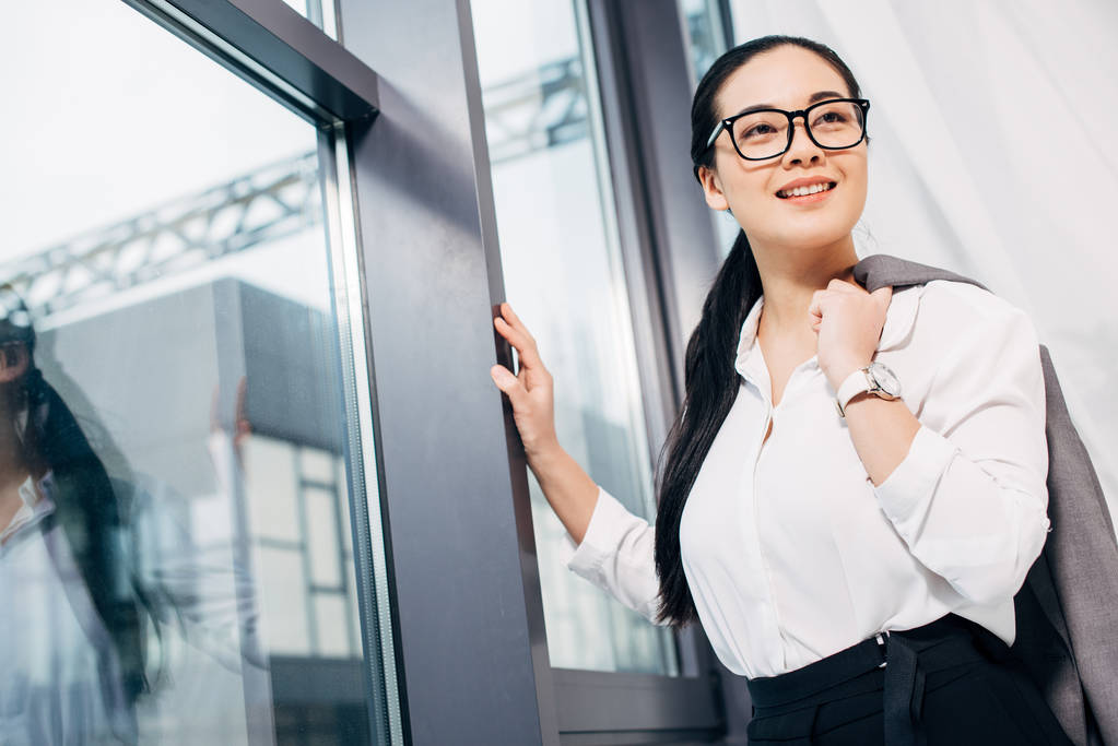 pretty businesswoman with jacket over shoulder standing by window in office - Photo, Image