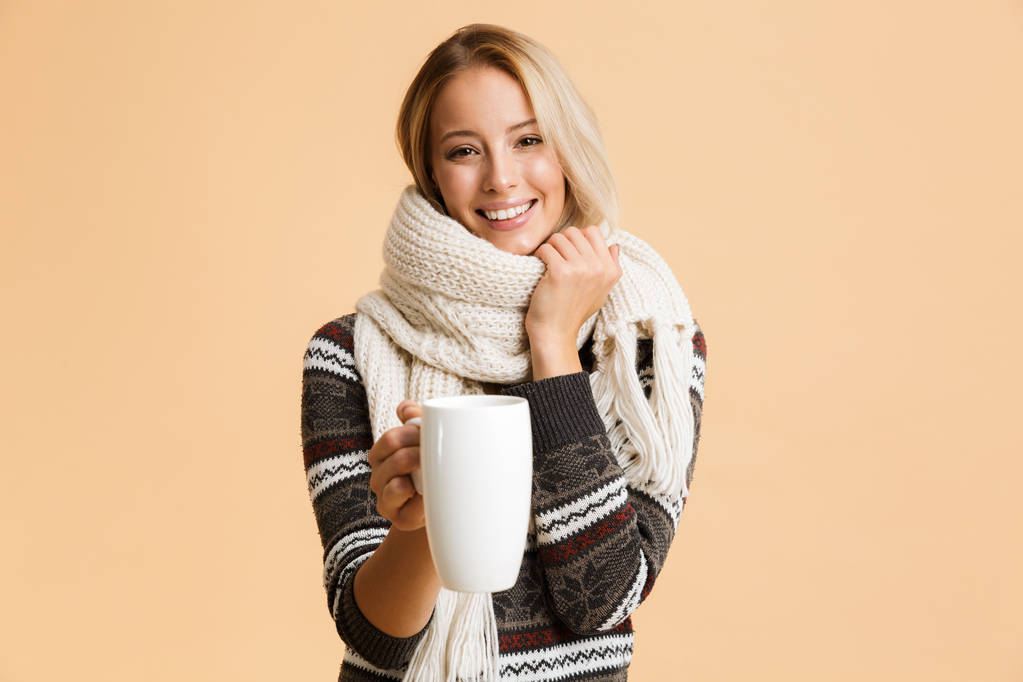 Портрет веселой девушки, одетой в свитер и шарф на бежевом фоне, с чашкой горячего чая
 - Фото, изображение
