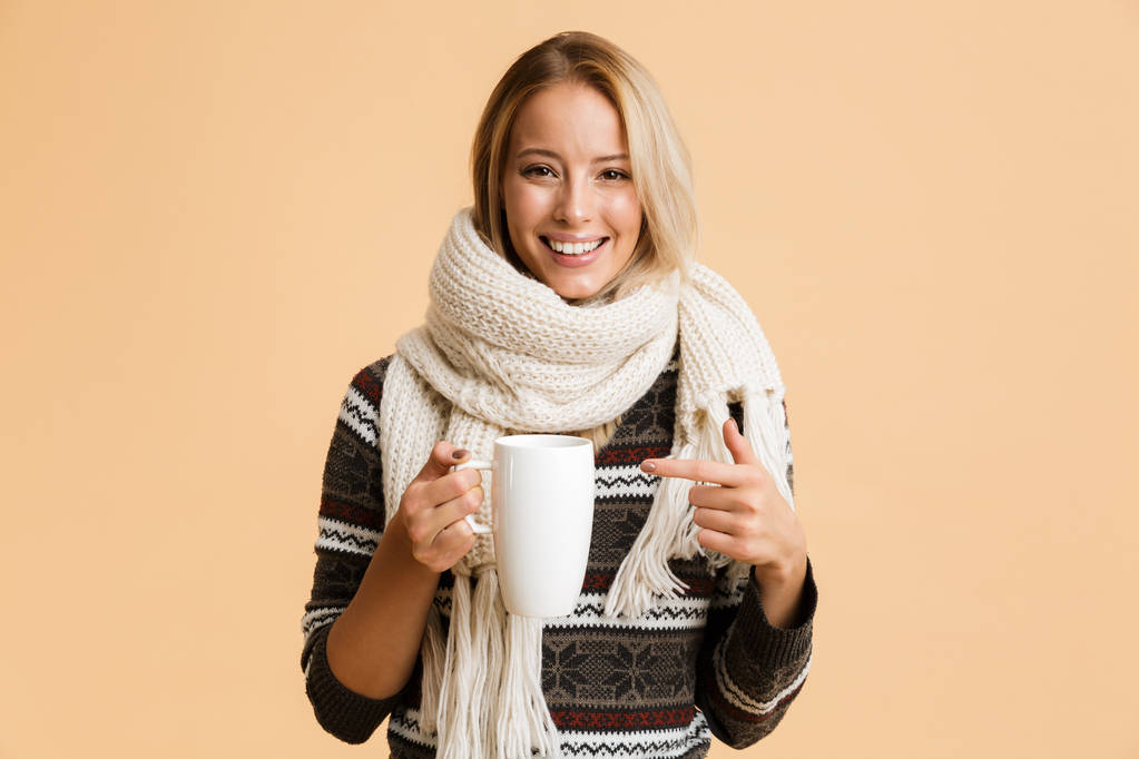 Portrait of a happy girl dressed in sweater and scarf isolated over beige background, holding cup of hot tea, pointing finger - Photo, Image