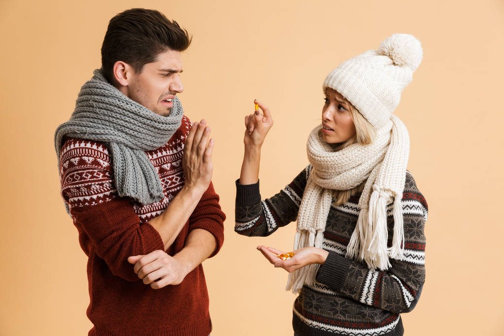 Portrait of a sick young couple dressed in sweaters and scarves standing together isolated over beige background, girl giving her boyfriend medication - Photo, Image