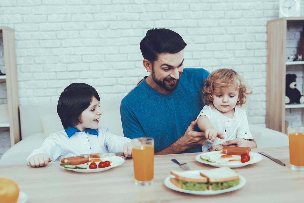 Ο άνθρωπος ξοδεύει το χρόνο με τους γιους του. Ο πατέρας των αγοριών συμμετέχει στην ανατροφή παιδιών. Ο πατέρας είναι σίτιση μια γιοι ένα πρωινό. Άνθρωποι είναι κάθεται στις καρέκλες στο τραπέζι. Άτομα που βρίσκονται στην κουζίνα. - Φωτογραφία, εικόνα