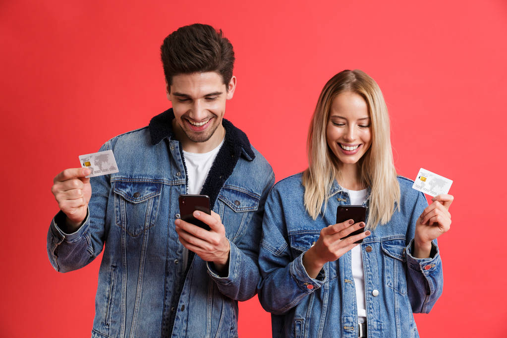 Muotokuva hymyilevä nuori pari pukeutunut denim takit seisoo yhdessä eristetty yli punainen tausta, matkapuhelimilla, osoittaa luottokortteja
 - Valokuva, kuva