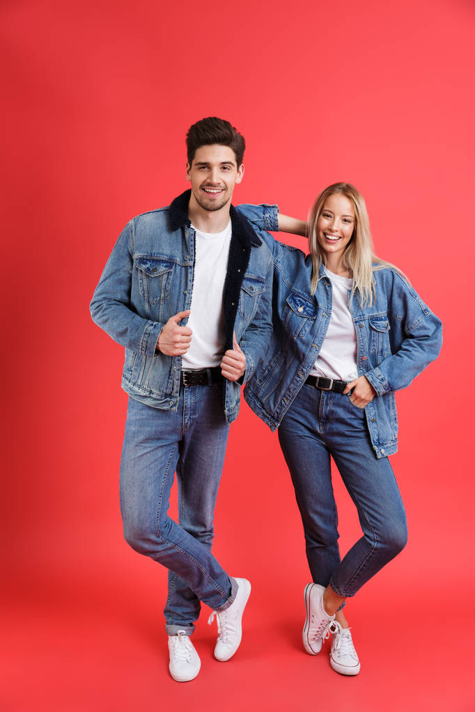 Full length portrait of a cheerful young couple dressed in denim jackets standing together isolated over red background - Photo, Image