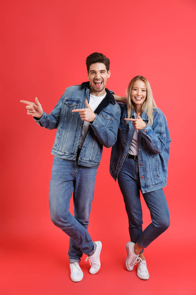 Портрет щасливої молодої пари, одягнений у джинсовий одяг, ізольований на червоному тлі, спрямовуючи пальці
 - Фото, зображення