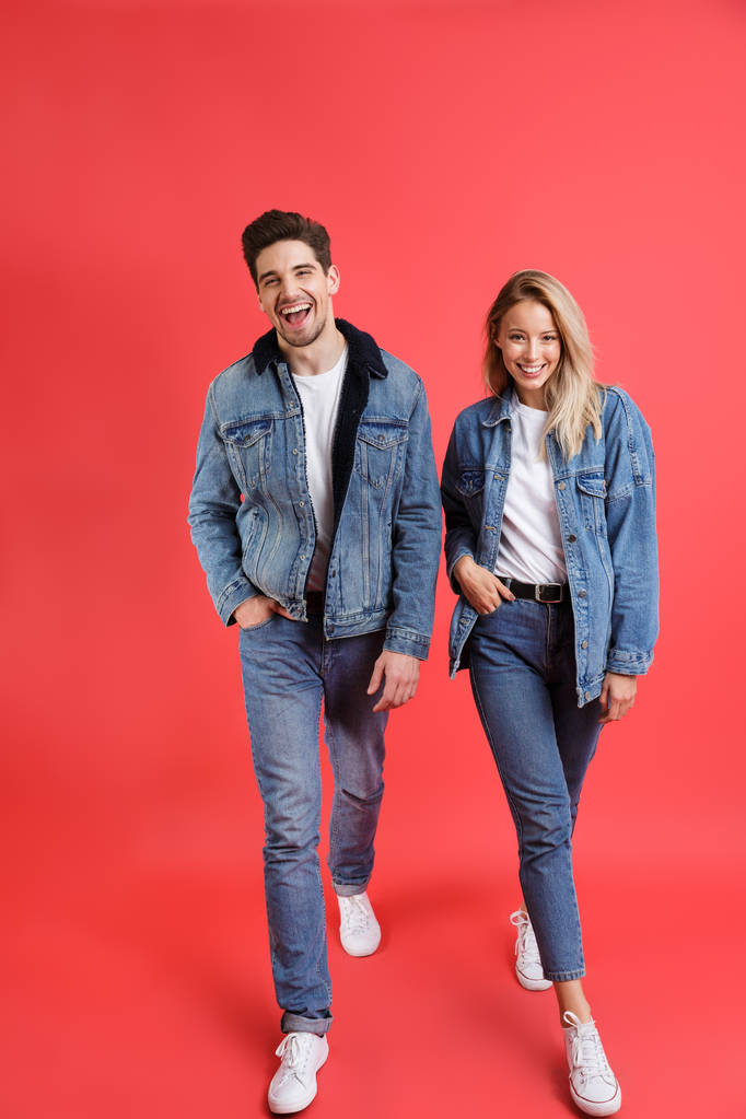 Full length portrait of a smiling young couple dressed in denim clothes isolated over red background, walking - Photo, Image