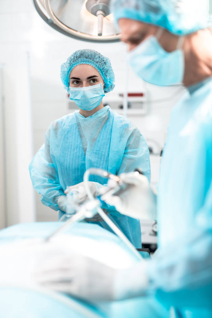 Verpleegkundige in blauwe jurk zuurstofmasker op patiënt gezicht houden tijdens chirurgie - Foto, afbeelding