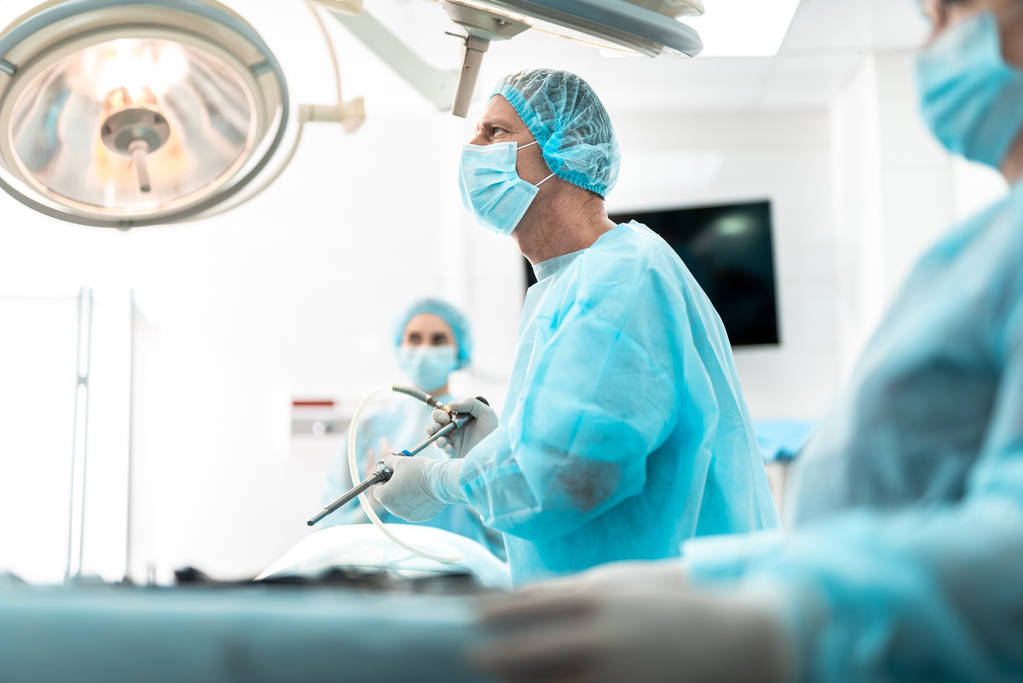 Chirurg in beschermend masker met behulp van de laparoscoop tijdens operatie - Foto, afbeelding