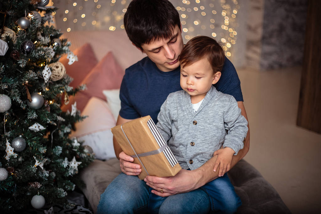 Petit garçon dans une veste décontractée et un jean assis près de l'arbre de Noël avec son père. Jeune homme et son fils
 - Photo, image