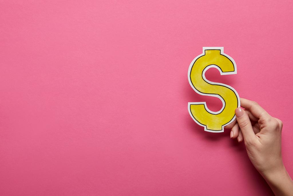 vista elevada da mão segurando sinal de dólar amarelo no fundo rosa
 - Foto, Imagem