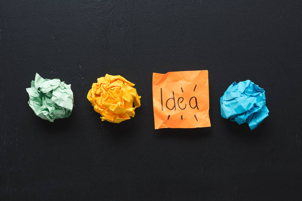Draufsicht auf "Idee" -Wort auf Haftnotiz mit bunten zerknüllten Papierkugeln auf schwarzem Hintergrund, Ideenkonzept - Foto, Bild