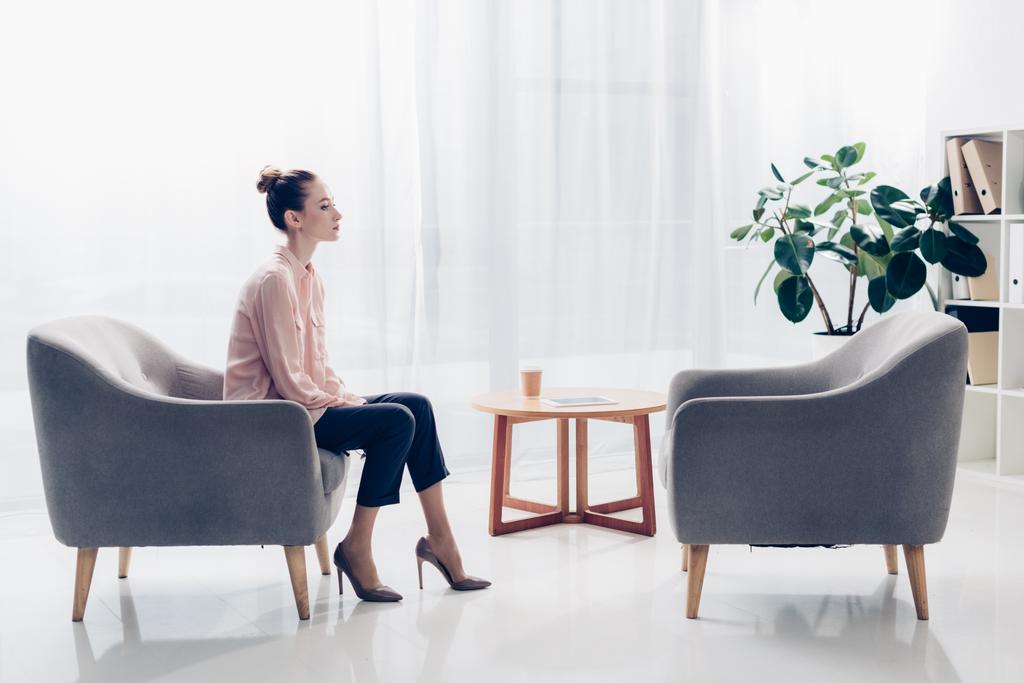 vista lateral de hermosa mujer de negocios sentada en sillón en la oficina y mirando hacia otro lado
 - Foto, Imagen