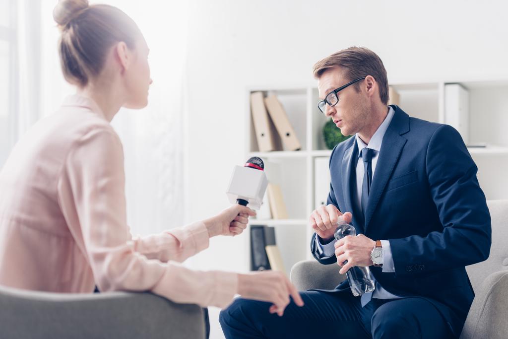 handsome businessman holding bottle of water and giving interview to journalist in office - Photo, Image