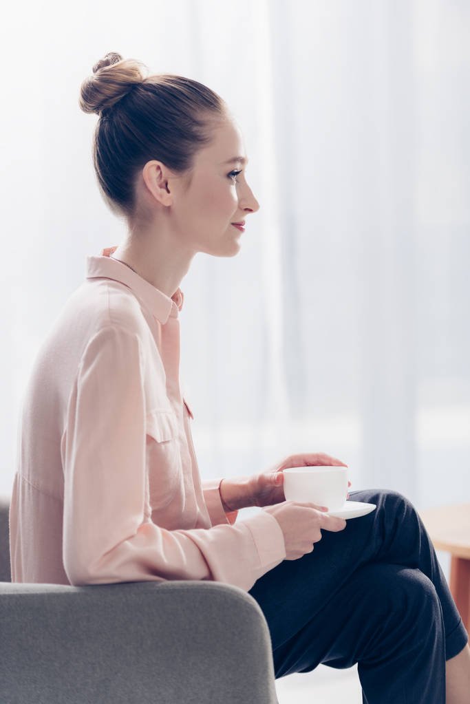 vista lateral de alegre hermosa mujer de negocios sentada con taza de té en sillón en la oficina
 - Foto, imagen