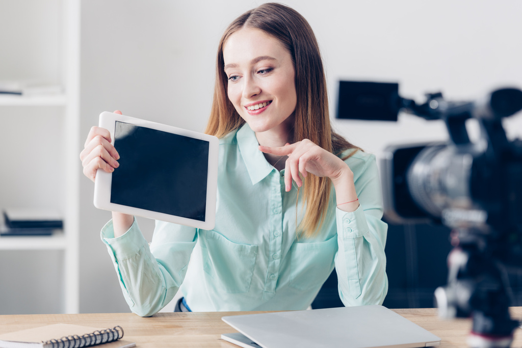 sonriente atractiva mujer video blogger grabación vlog y apuntando en la tableta con pantalla en blanco en la oficina
 - Foto, imagen