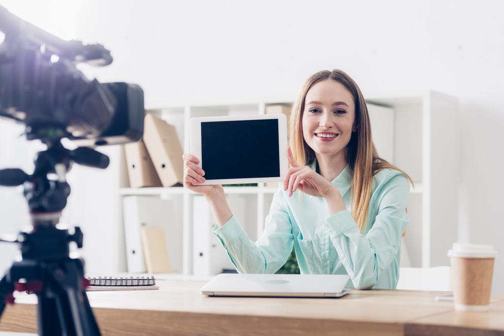 smiling attractive female video blogger recording vlog and showing tablet with blank screen in office - Photo, Image
