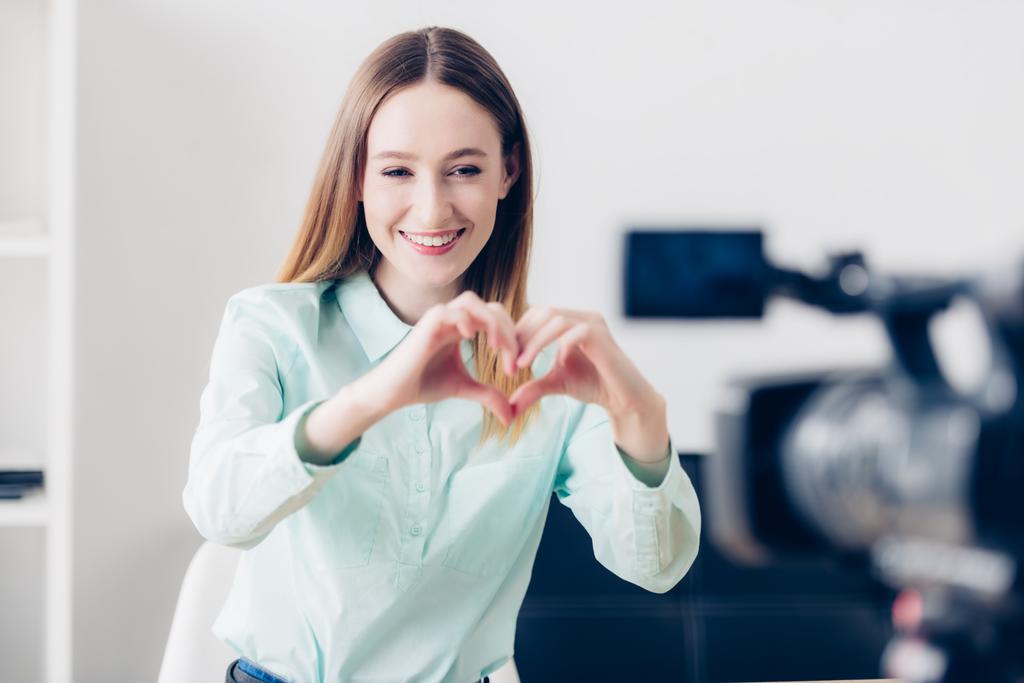 Gelukkig aantrekkelijke vrouwelijke video blogger opname vlog en tonen hart met vingers in office - Foto, afbeelding