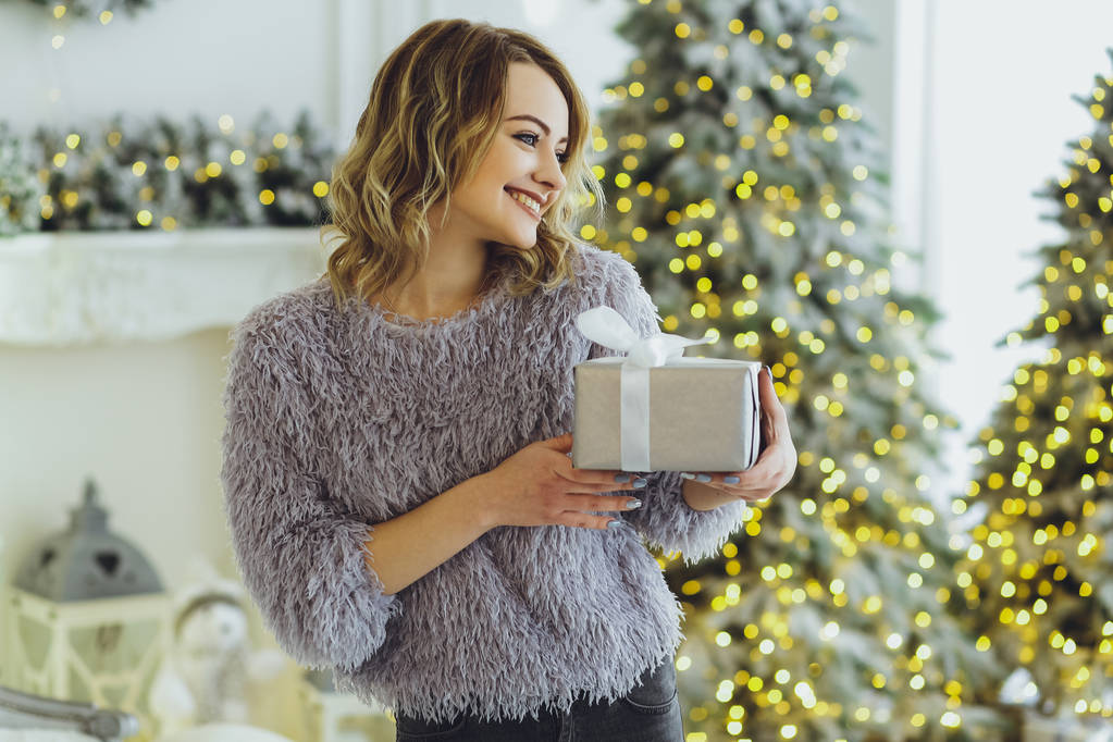 Молодая красивая блондинка в сером свитере с подарком на руках возле Нового года, елка с подарками
 - Фото, изображение