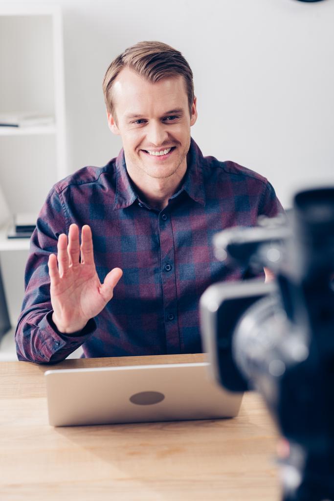 χαμογελώντας όμορφος αρσενικό βίντεο blogger καταγραφή vlog και κουνώντας το χέρι του στο γραφείο - Φωτογραφία, εικόνα