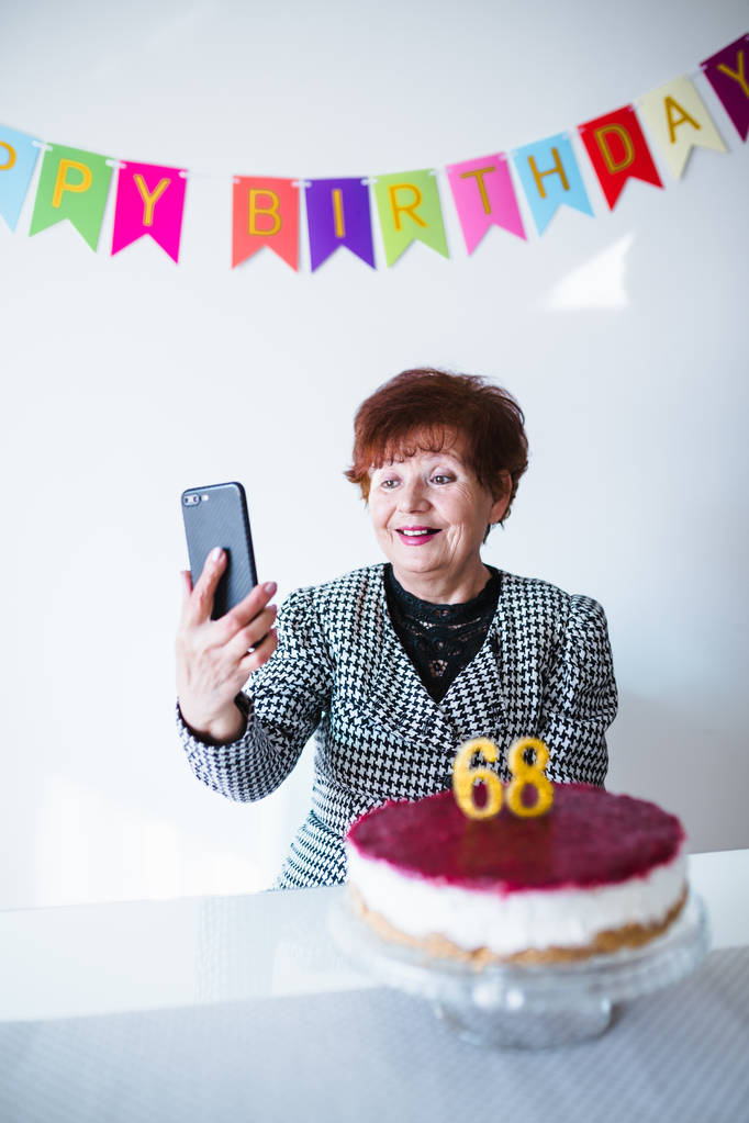 Donna anziana che fa selfie vicino al tavolo con la torta mentre festeggia il compleanno a casa
 - Foto, immagini