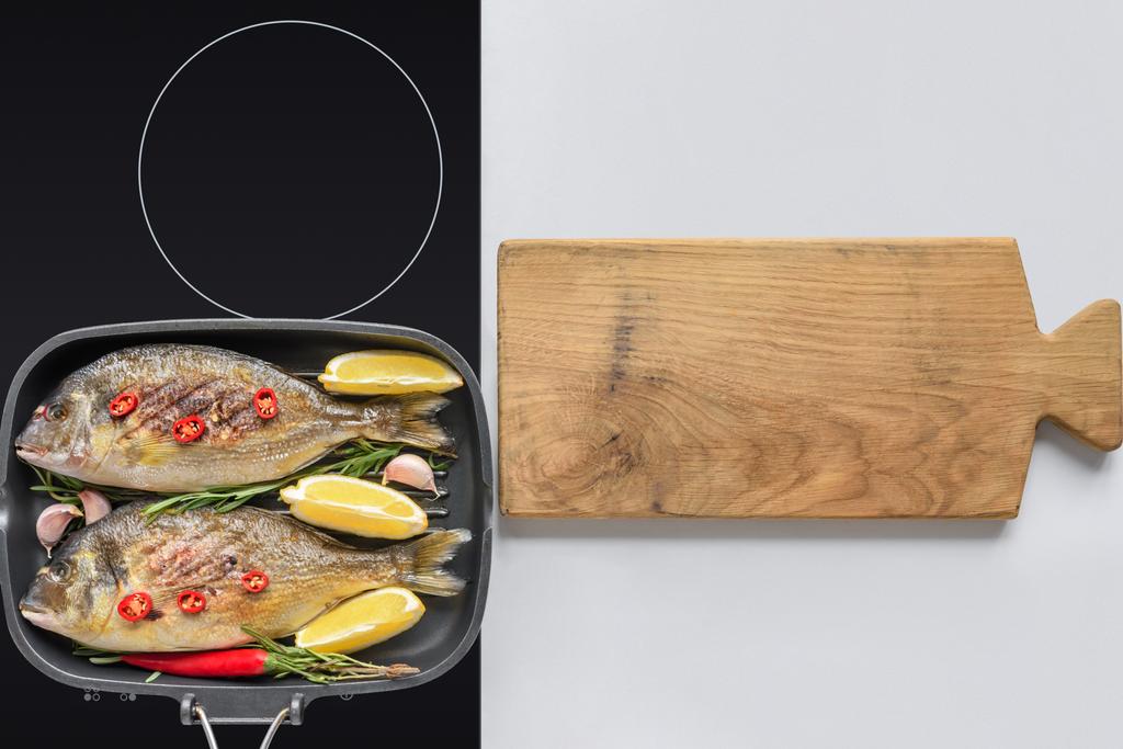 bovenaanzicht van heerlijke gebakken vis op elektrisch fornuis en houten snijplank - Foto, afbeelding