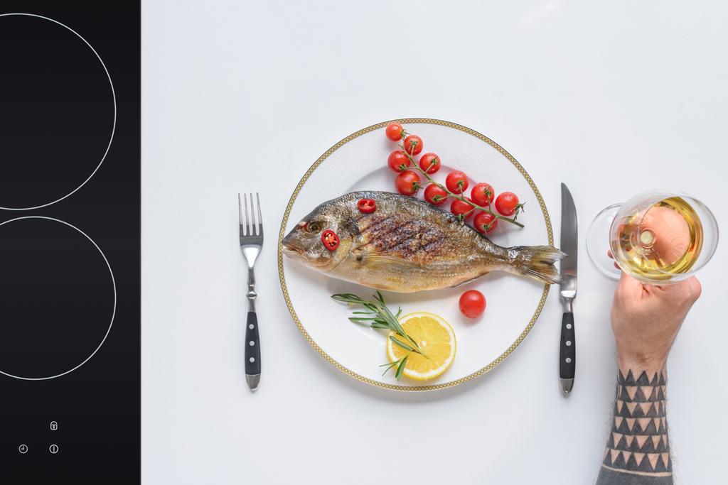 bijgesneden schot van houder glas wijn en heerlijke gebakken vis op plaat - Foto, afbeelding