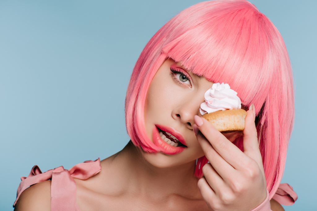 sinnliche junge Frau in rosa Perücke posiert mit leckeren Cupcake isoliert auf blau - Foto, Bild