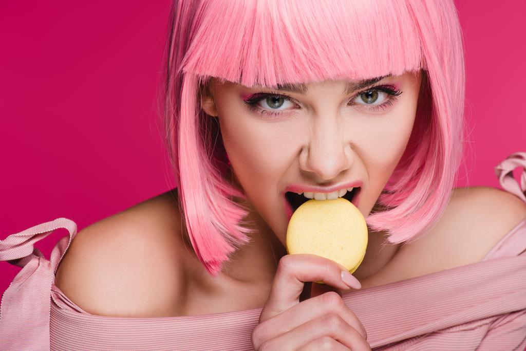 attraktive aggressive Mädchen in rosa Perücke beißt Macaron isoliert auf rosa - Foto, Bild