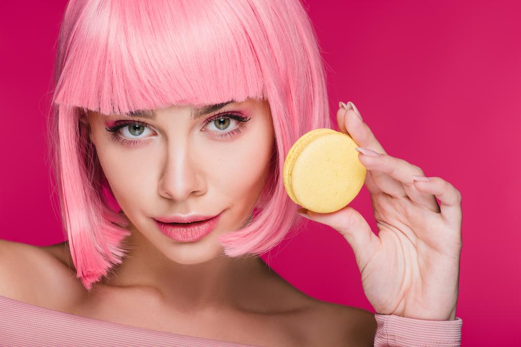 attractive young woman in pink wig posing with yellow macaron isolated on pink - Photo, Image