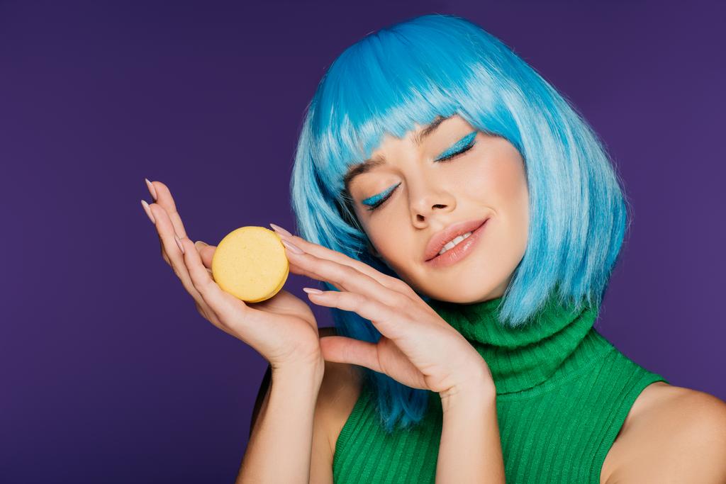 verträumtes Mädchen in blauer Perücke posiert mit Macaron isoliert auf lila - Foto, Bild