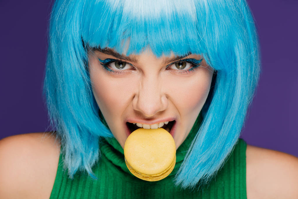 agresivní mladá žena v modrá paruka kousání macaron izolované na fialová - Fotografie, Obrázek