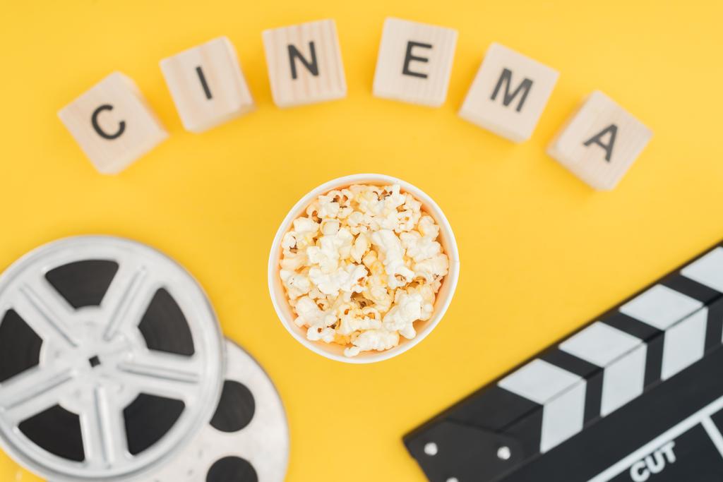 Ansicht von Holzwürfeln mit "Kino" -Schriftzug, Klappbrett, Filmrollen und Popcorneimer isoliert auf gelb - Foto, Bild