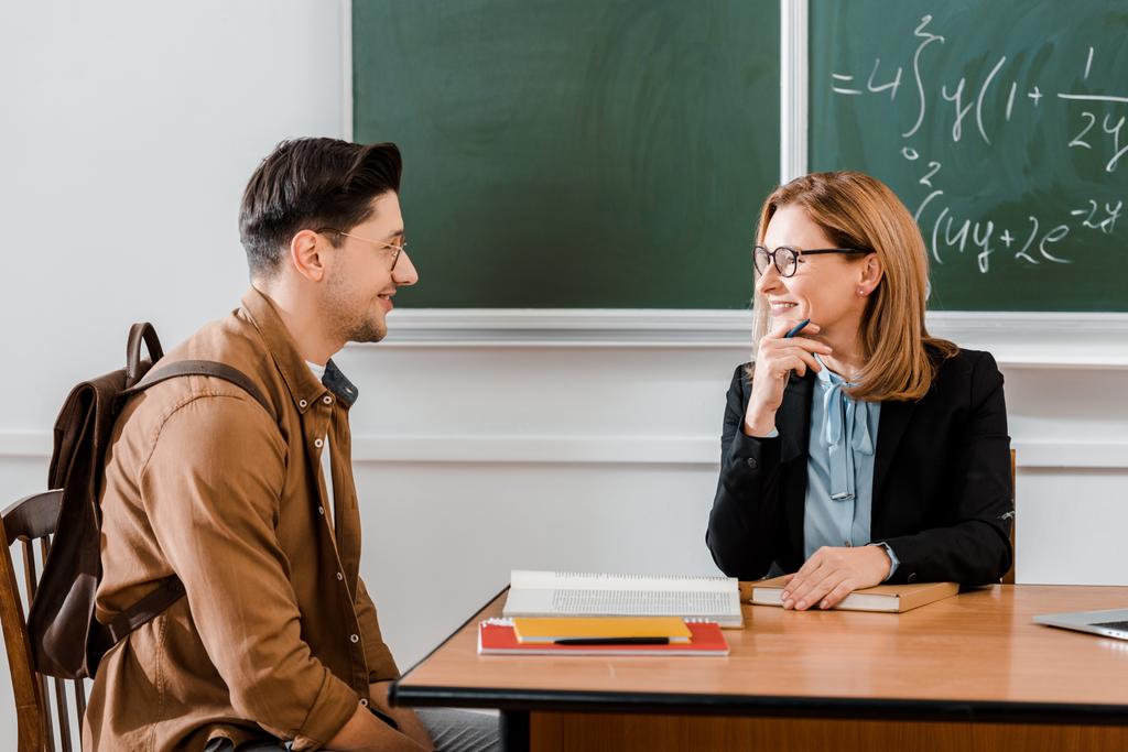 Жінка-вчителька посміхається біля молодого студента з рюкзаком в класі
  - Фото, зображення