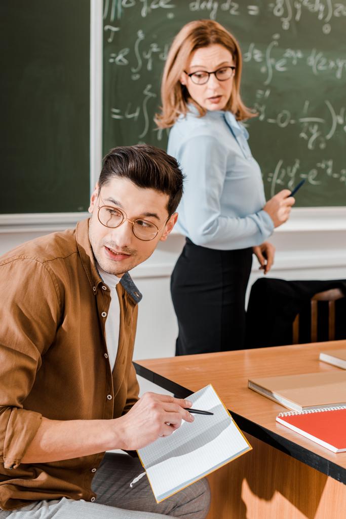 Jovem estudante olhando para longe e segurando notebook com caneta perto do professor feminino
  - Foto, Imagem