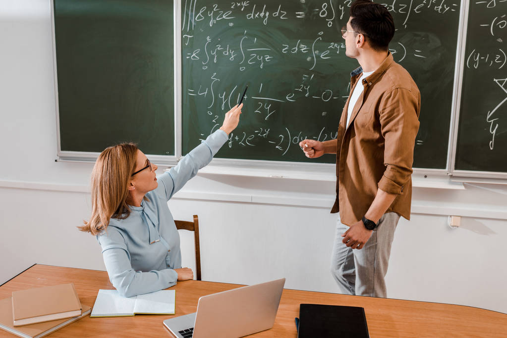 Вчителька вказує на рівняння, коли студент стоїть біля дошки
  - Фото, зображення