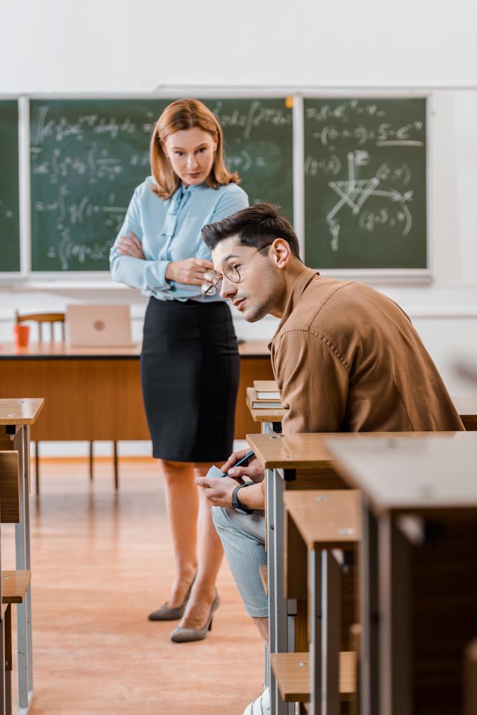 enseignante insatisfaite avec les bras croisés regardant étudiant mâle tricher pendant l'examen en classe
 - Photo, image