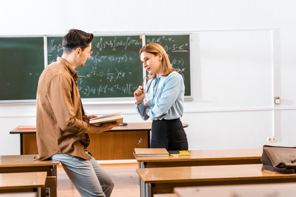 Estudante masculino segurando livro perto do professor feminino durante a aula em sala de aula
 - Foto, Imagem