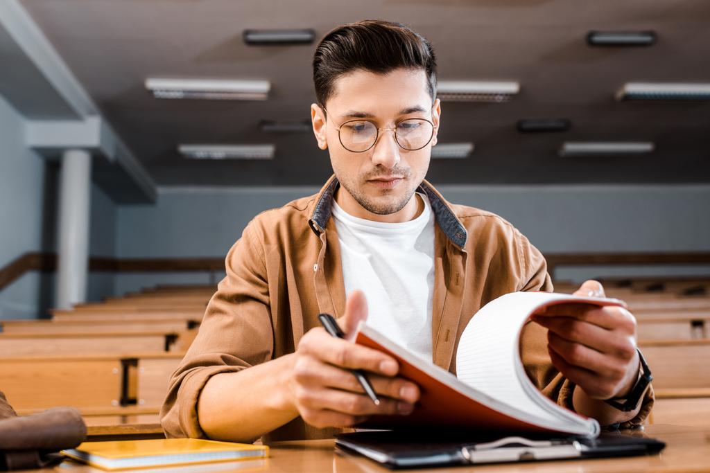 koncentrált férfi hallgató, szemüveg, ülő-on iskolapad és notebook tartó tollal a tanterem - Fotó, kép