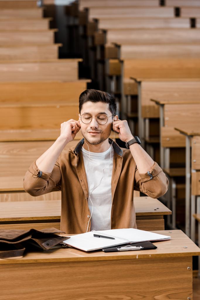männlicher Student mit Brille sitzt am Schreibtisch und setzt sich im Hörsaal Kopfhörer auf - Foto, Bild