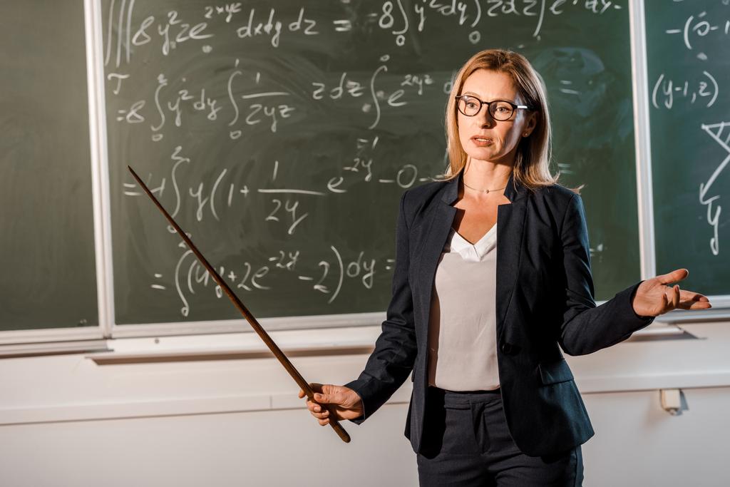 учительница в формальной одежде с деревянным указателем, объясняющим математические уравнения в классе
 - Фото, изображение