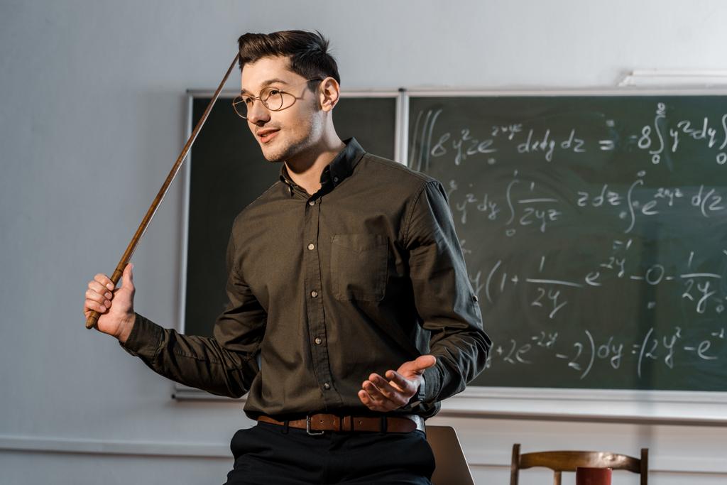 guapo maestro masculino en ropa formal sosteniendo puntero de madera y explicando ecuaciones en clase
 - Foto, Imagen