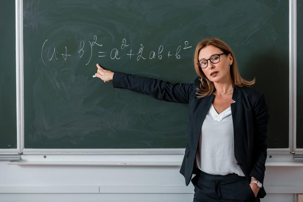 vrouwelijke leraar camera kijken en wijzen met de vinger naar wiskundige vergelijking op een schoolbord in de klas - Foto, afbeelding