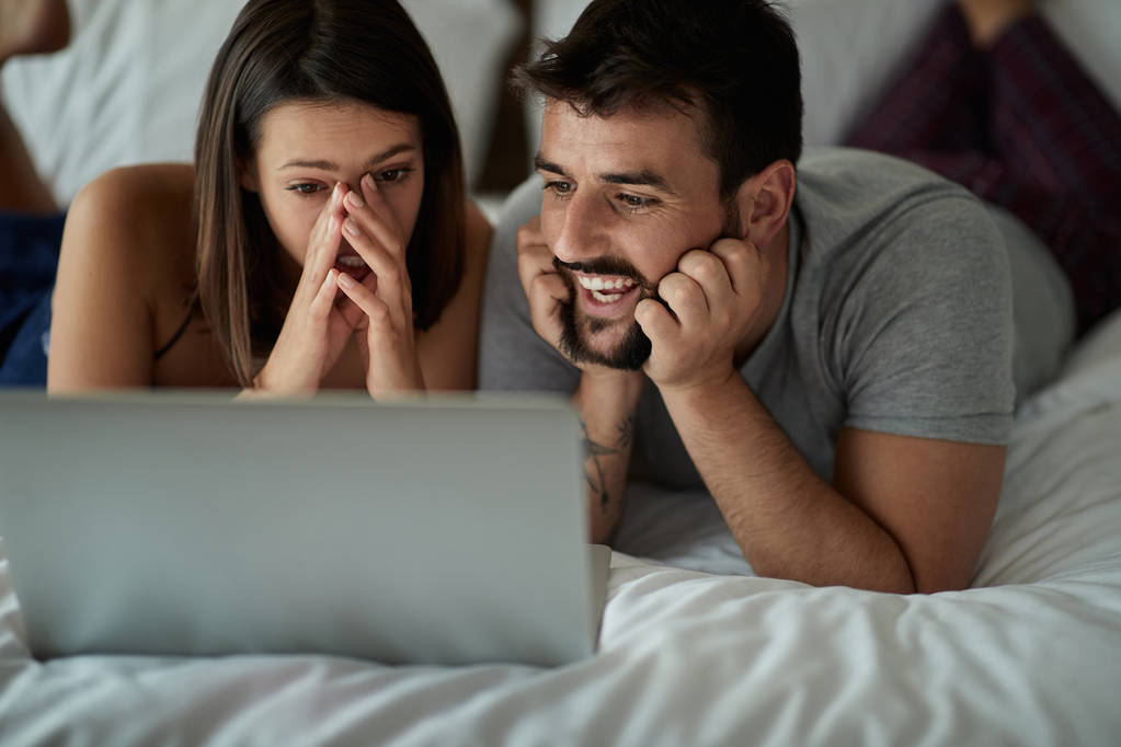 Laughing boyfriend and girlfriend in bedroom with laptop watching movie in bed - Photo, Image
